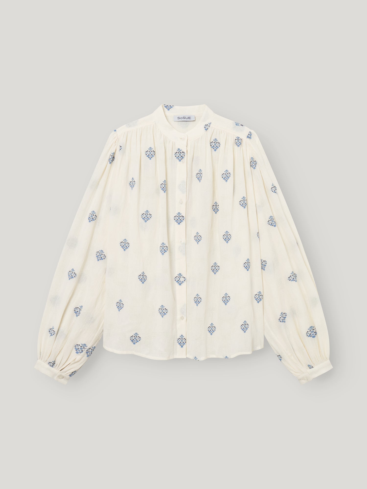 Die Bluse Embroidery ist eine weiße Damenbluse mit langen Ärmeln, einem hohen Kragen, plissierter Vorderseite, blauer geometrischer Blumenstickerei, geknöpften Manschetten und einer entspannten Passform für mühelosen Stil.