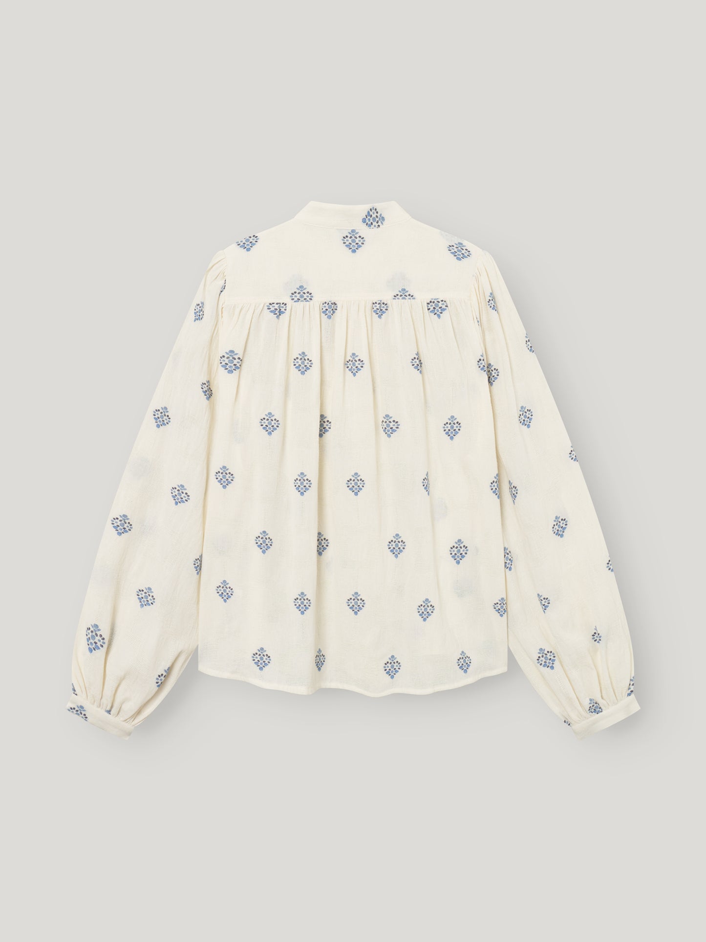 Die Bluse Embroidery ist eine cremefarbene langärmelige Damenbluse mit blauen geometrischen Mustern, blauer Stickerei, gerafften Passelementen und leicht gepufften Ärmeln. Die Abbildung zeigt die Bluse flach von hinten vor einem hellen Hintergrund.