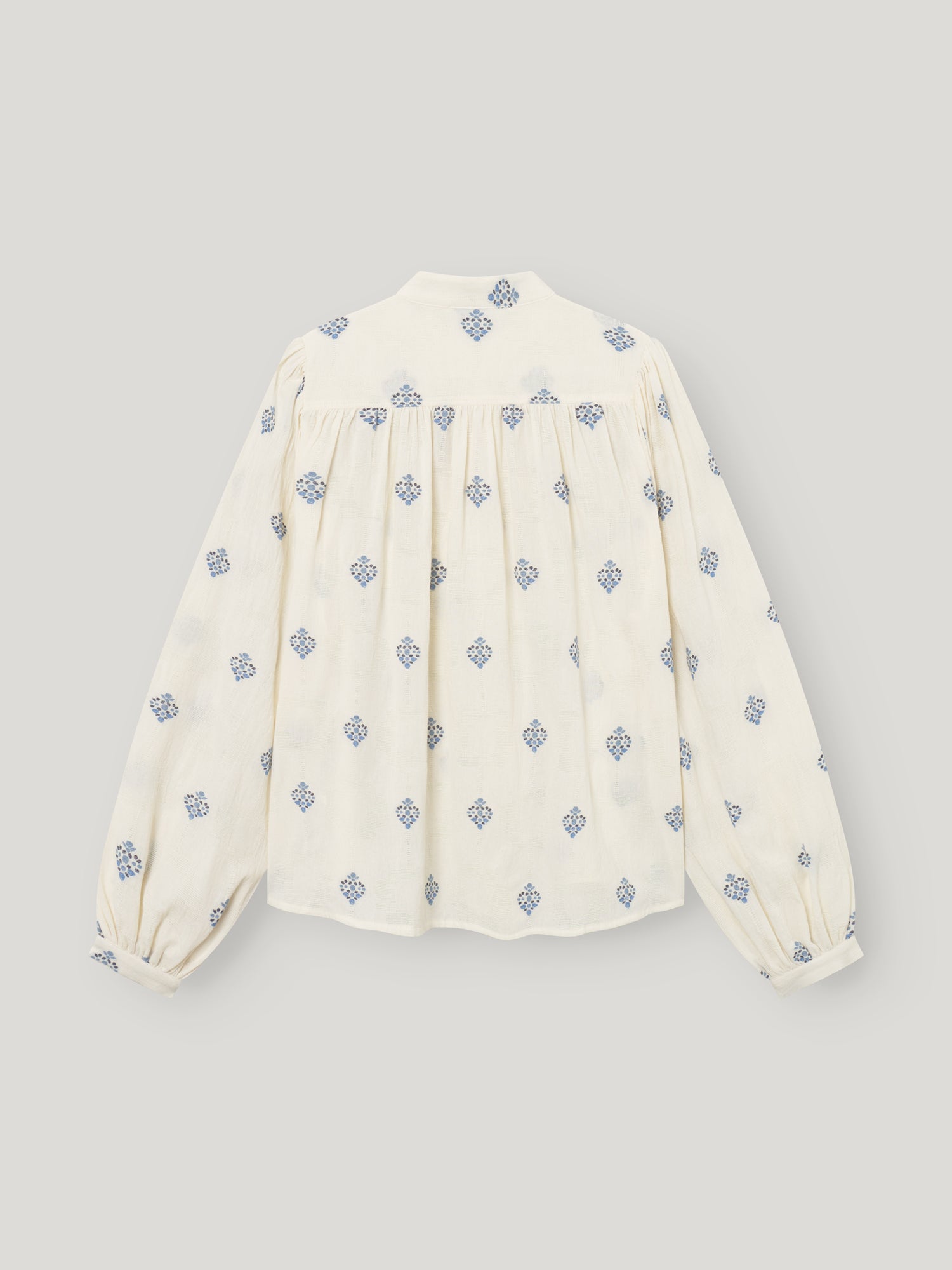 Die Bluse Embroidery ist eine cremefarbene langärmelige Damenbluse mit blauen geometrischen Mustern, blauer Stickerei, gerafften Passelementen und leicht gepufften Ärmeln. Die Abbildung zeigt die Bluse flach von hinten vor einem hellen Hintergrund.
