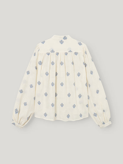 Die Bluse Embroidery ist eine cremefarbene langärmelige Damenbluse mit blauen geometrischen Mustern, blauer Stickerei, gerafften Passelementen und leicht gepufften Ärmeln. Die Abbildung zeigt die Bluse flach von hinten vor einem hellen Hintergrund.
