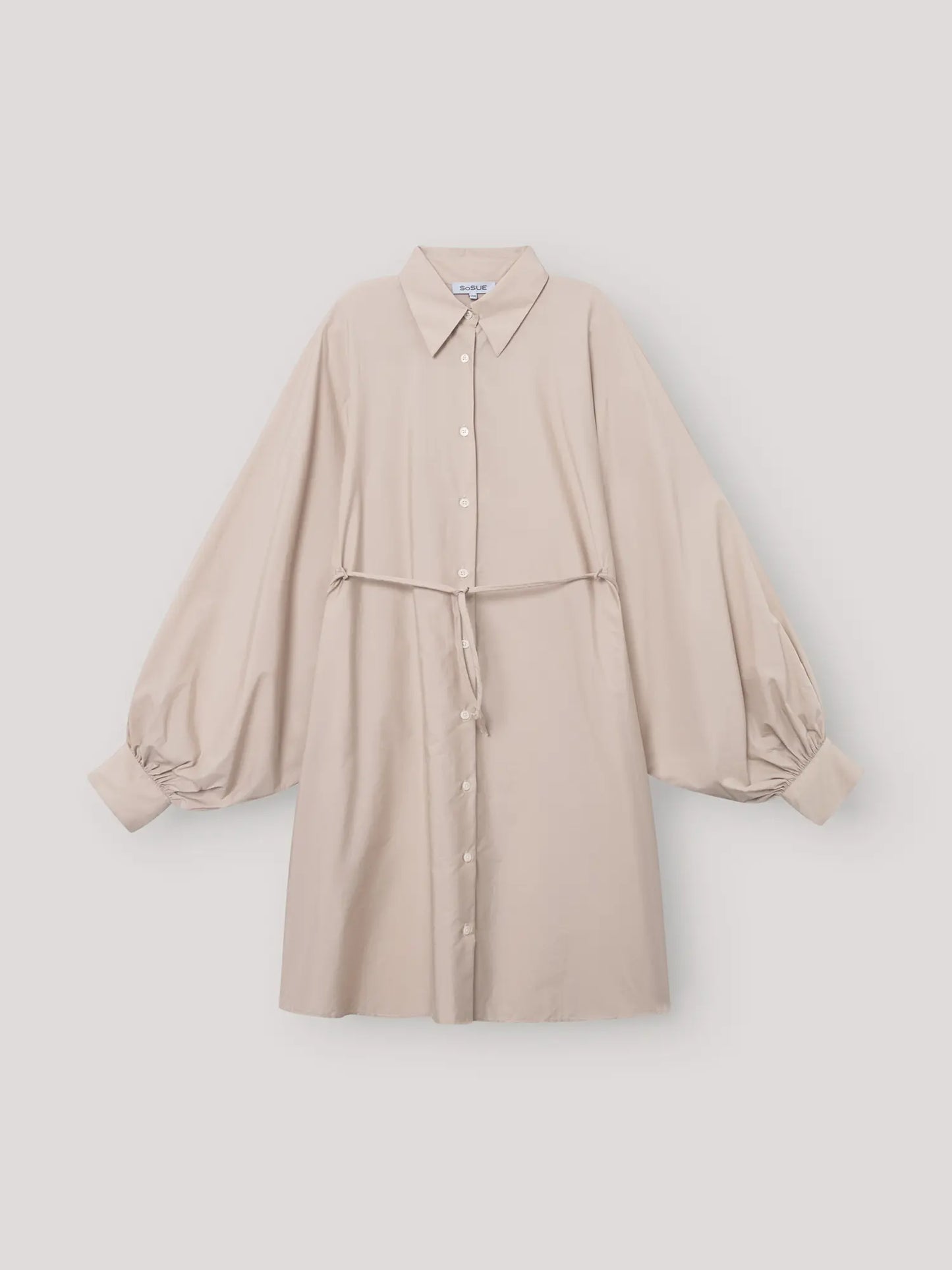 Das Kleid Antonia Dress Vol. 2 Beige ist ein beigefarbenes Hemdkleid mit Kragen und Knopfleiste, langen Puffärmeln, gerafften Manschetten und einem schmalen, passenden Gürtel in der Taille, das vor einem schlichten, hellen Hintergrund gezeigt wird.