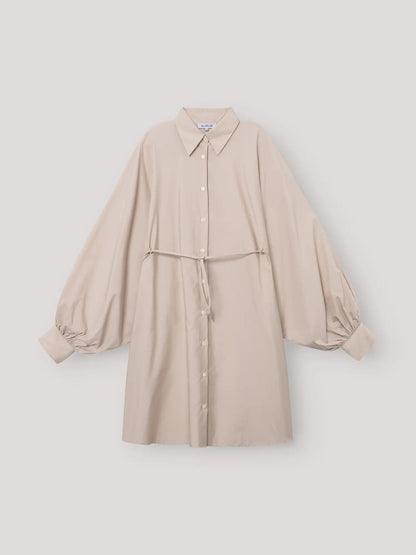 Das Kleid Antonia Dress Vol. 2 Beige ist ein beigefarbenes Hemdkleid mit Kragen und Knopfleiste, langen Puffärmeln, gerafften Manschetten und einem schmalen, passenden Gürtel in der Taille, das vor einem schlichten, hellen Hintergrund gezeigt wird.