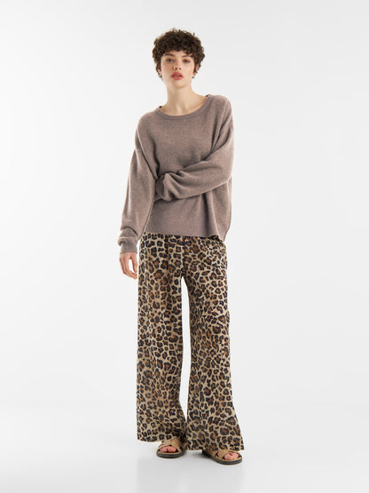 Eine Person mit kurzen lockigen Haaren trägt einen lockeren Pullover Boyfriend azteco, eine Hose mit weitem Bein und Leopardenmuster sowie Sandalen und steht vor einem schlichten weißen Hintergrund.