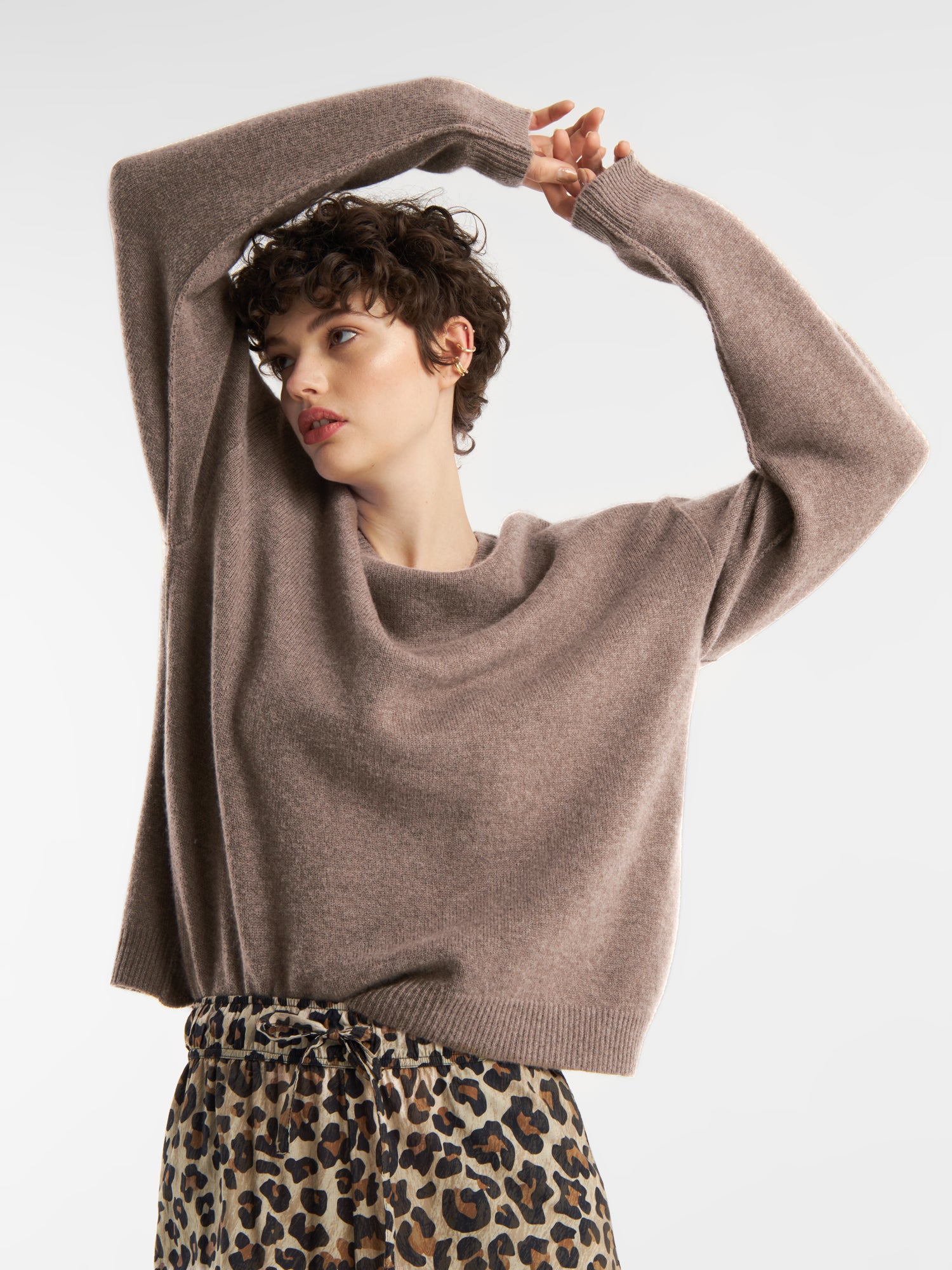 Eine Person mit kurzen, lockigen Haaren trägt einen lockeren Pullover Boyfriend azteco mit einer hochtaillierten Hose mit Leopardenmuster und posiert mit erhobenen Armen und Blick zur Seite vor einem schlichten weißen Hintergrund.