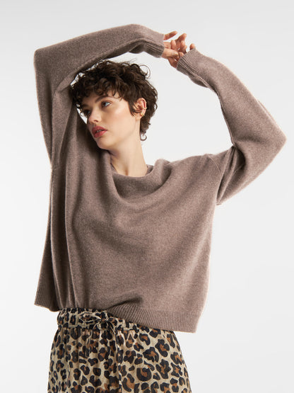 Eine Person mit kurzen, lockigen Haaren trägt einen lockeren Pullover Boyfriend azteco mit einer hochtaillierten Hose mit Leopardenmuster und posiert mit erhobenen Armen und Blick zur Seite vor einem schlichten weißen Hintergrund.