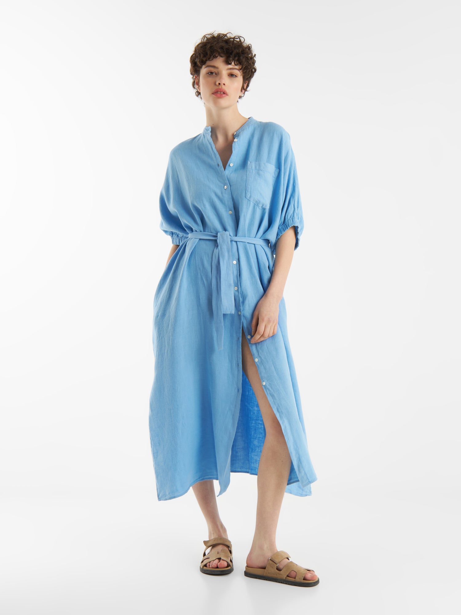 Eine Person mit kurzen lockigen Haaren trägt das Kleid Cowboy Button Linen Blue, ein locker sitzendes, gegürtetes Hemdkleid mit gekrempelten Ärmeln und einem Schlitz an der Vorderseite, gepaart mit hellbraunen Sandalen, vor einem schlichten weißen Hintergrund.