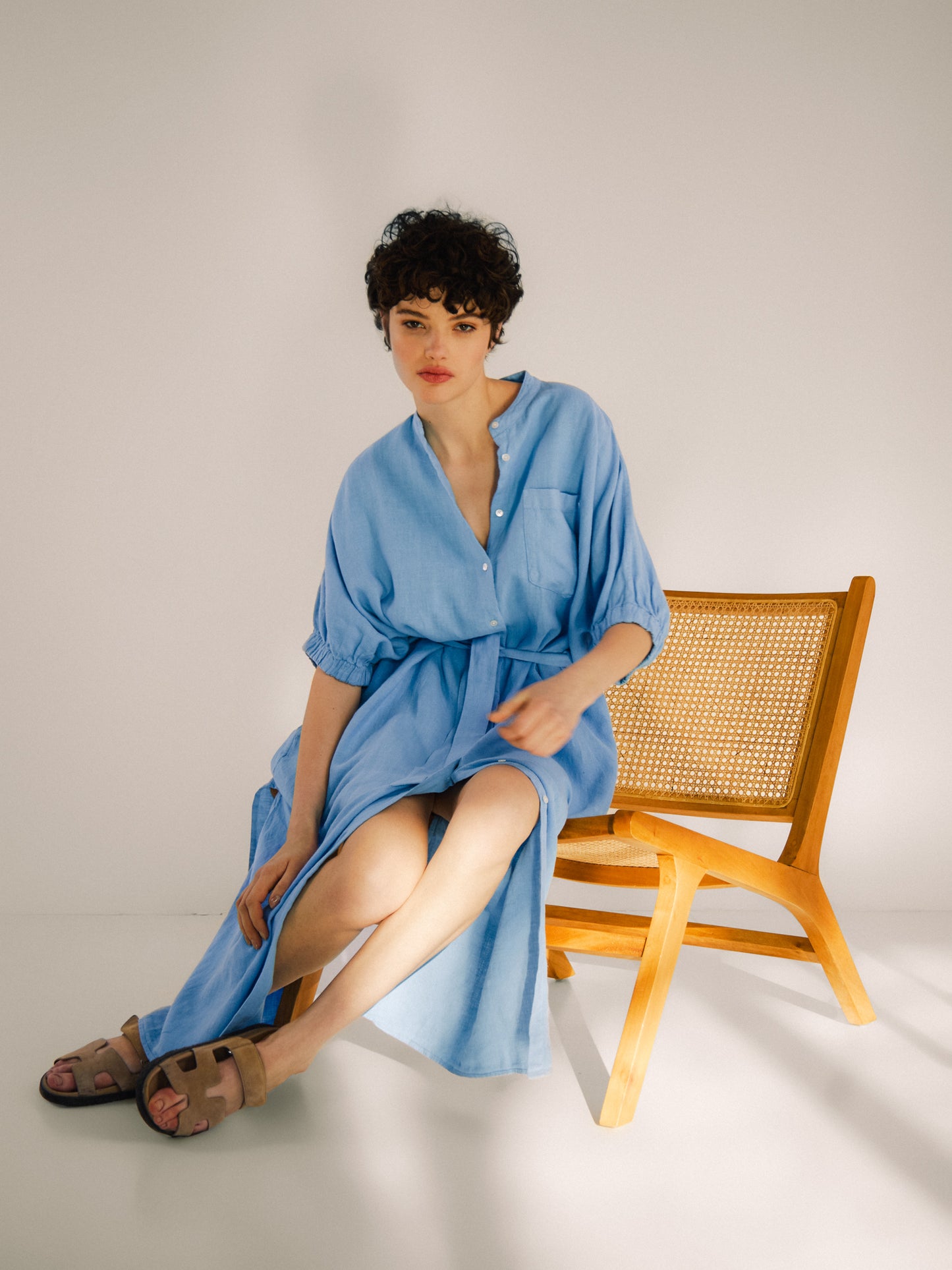 Eine Frau mit kurzen lockigen Haaren in einem hellblauen Kleid Cowboy Button Linen sitzt auf der Kante eines Holzstuhls in einem hellen, minimalistischen Raum und trägt beigefarbene Sandalen.