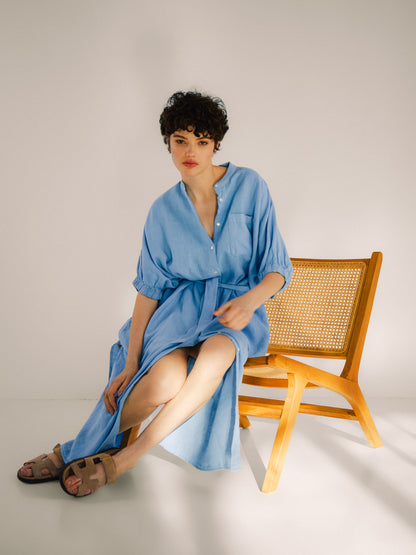 Eine Frau mit kurzen lockigen Haaren in einem hellblauen Kleid Cowboy Button Linen sitzt auf der Kante eines Holzstuhls in einem hellen, minimalistischen Raum und trägt beigefarbene Sandalen.