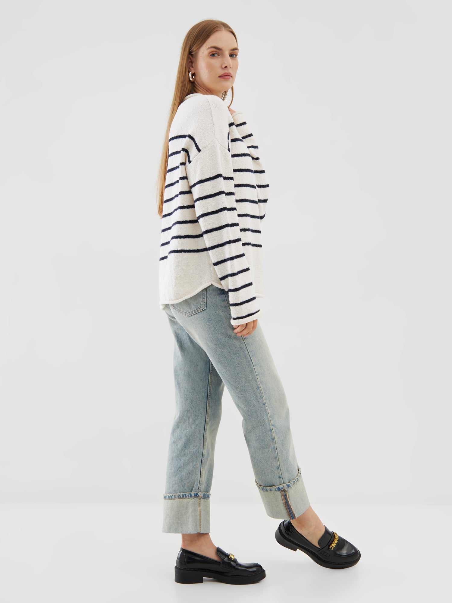 Eine Person mit langen, glatten Haaren trägt einen Pullover Sylt aus Bio-Baumwolle, hellblaue Jeans mit Bündchen und schwarze Halbschuhe. Sie steht seitlich vor einem schlichten, hellen Hintergrund.