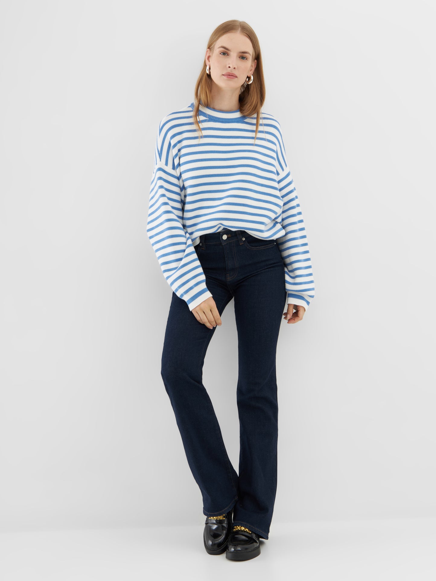 Eine Frau steht vor einem schlichten weißen Hintergrund und trägt den Pullover Sailor Vol. 3, dunkelblaue Jeans und schwarze Slipper mit goldenen Kettendetails. Sie hat glattes hellbraunes Haar und blickt direkt in die Kamera.