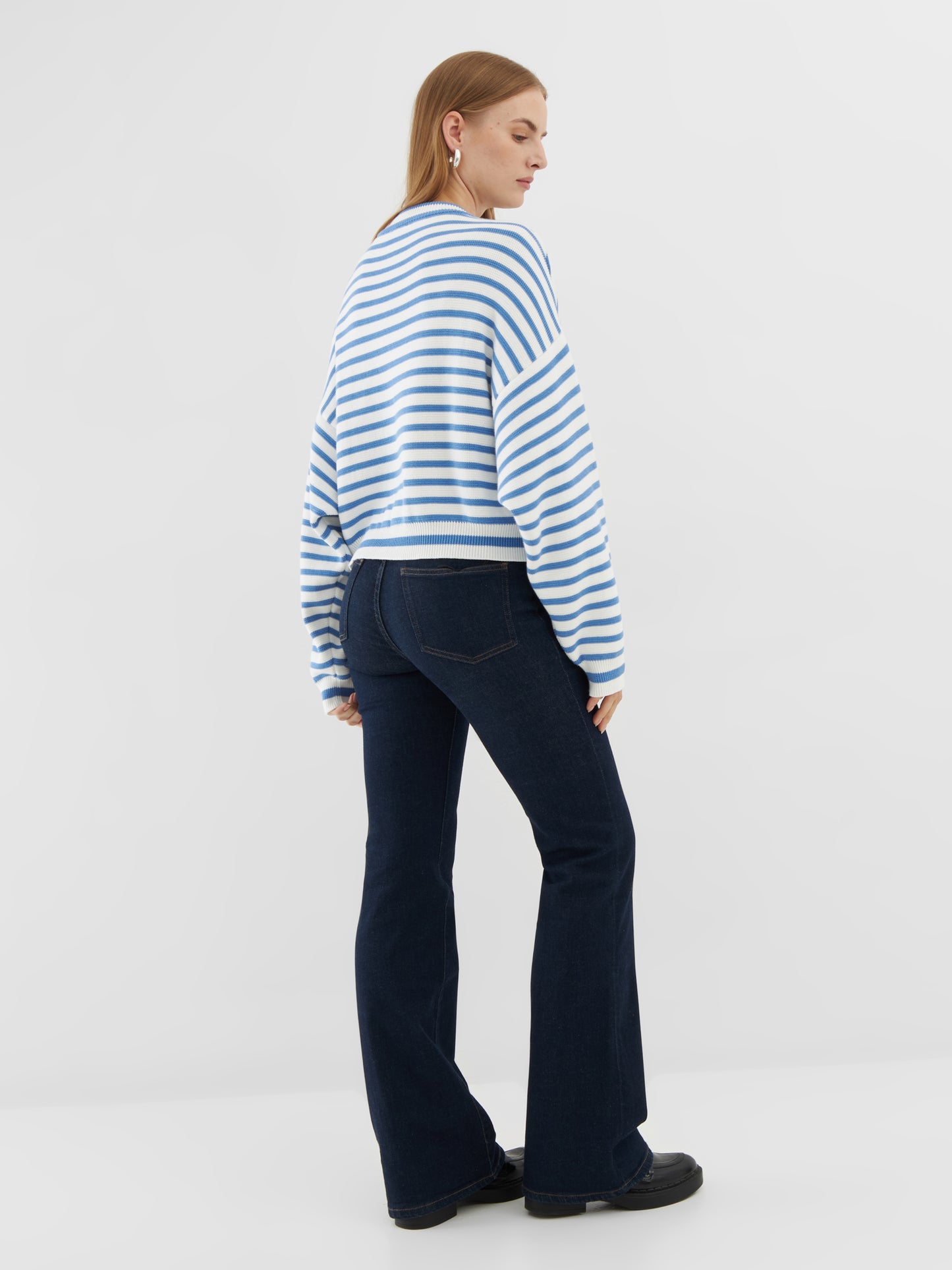 Eine Frau blickt von sich weg und trägt den Pullover Sailor Vol. 3 im blau-weißen maritimen Stil, gepaart mit dunklen Bootcut-Jeans und schwarzen Schuhen vor einem schlichten weißen Hintergrund.