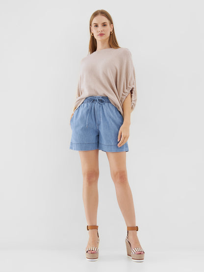 Eine Frau steht vor einem schlichten weißen Hintergrund und trägt das Top Cowboy Knit Beige, hellblaue Shorts mit Kordelzug und braune Plateau-Keilsandalen. Sie hat glattes Haar, die Hände in den Taschen und einen neutralen Gesichtsausdruck.