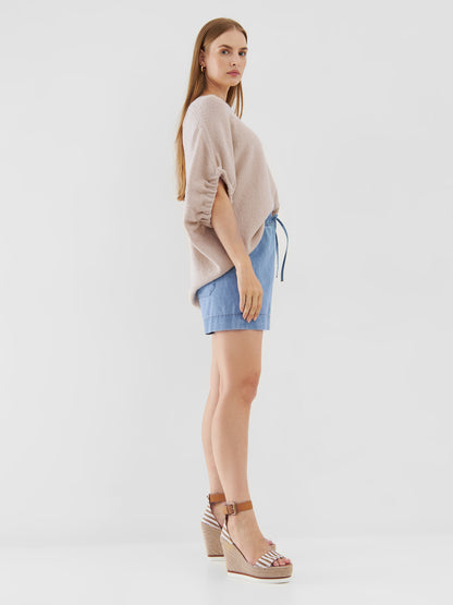 Eine Frau mit langem glattem Haar und neutralem Gesichtsausdruck steht seitlich vor einem schlichten weißen Hintergrund. Sie trägt das übergroße Top Cowboy Knit Beige, hellblaue Shorts und braune Plateausandalen mit Knöchelriemen.