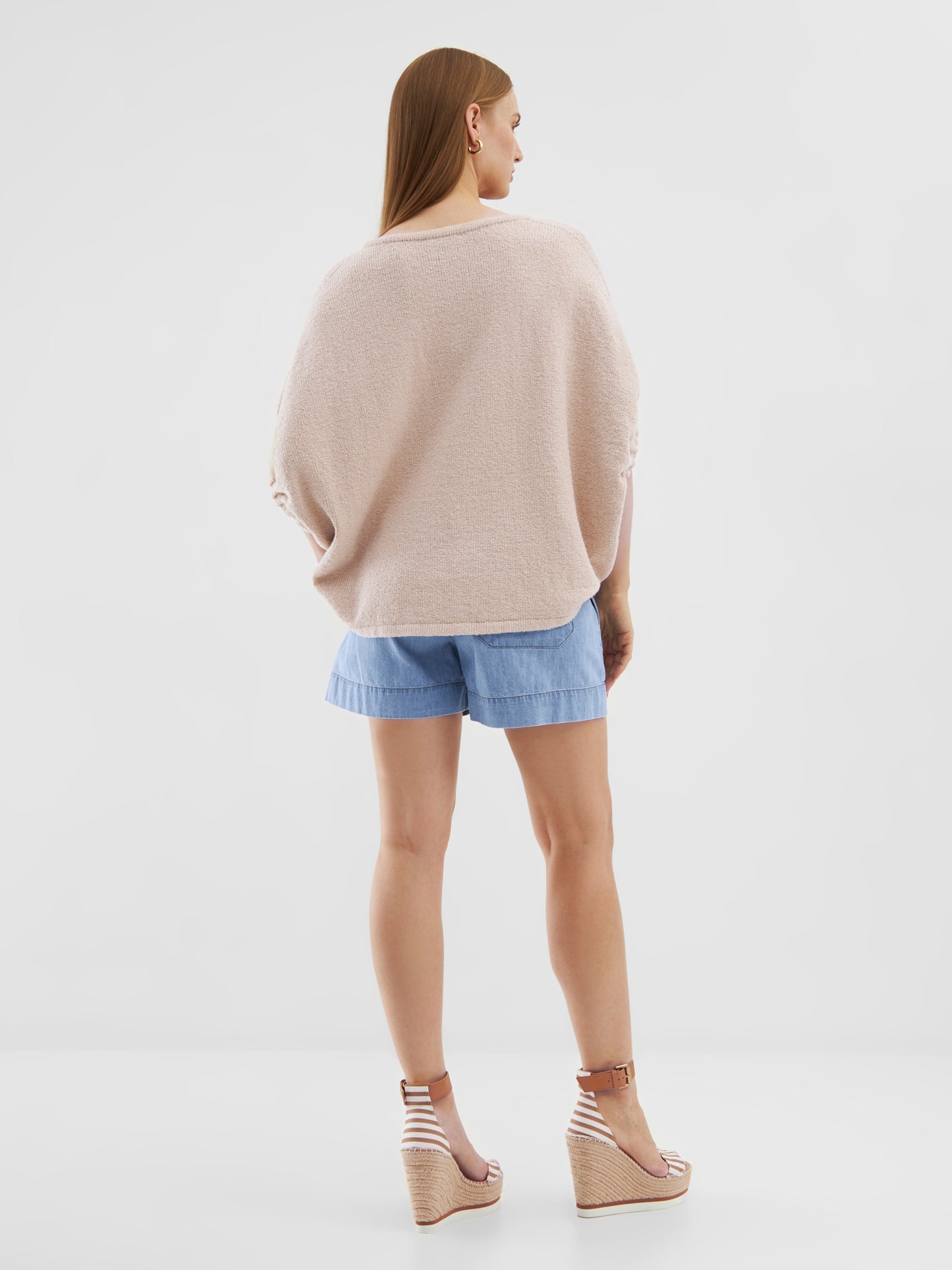 Eine Frau mit glattem hellbraunem Haar blickt von sich weg und trägt das übergroße Top Cowboy Knit Beige, hellblaue Shorts und beigefarbene Keilsandalen vor einem schlichten weißen Hintergrund.