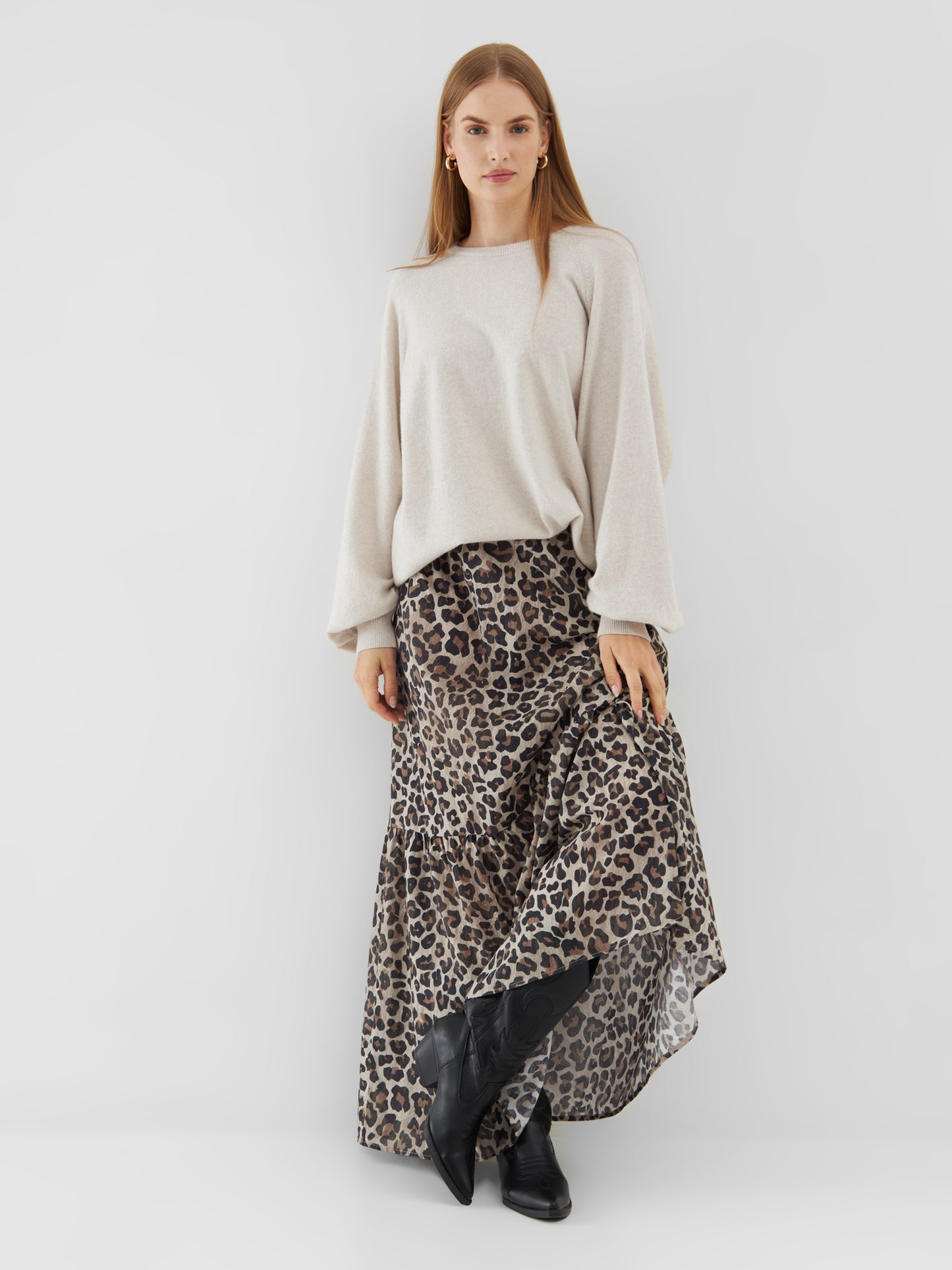 Eine Frau mit glattem, hellbraunem Haar und neutralem Gesichtsausdruck steht vor einem schlichten Hintergrund und trägt den Pullover Elli Beige, einen langen Stufenrock mit Leopardenmuster und schwarze Stiefel.