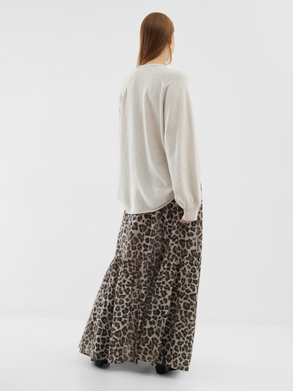 Eine Person mit langen Haaren blickt in die Ferne. Sie trägt den Pullover Elli Beige und einen langen, gestuften Rock mit Leopardenmuster und schwarzen Stiefeln vor einem schlichten weißen Hintergrund.