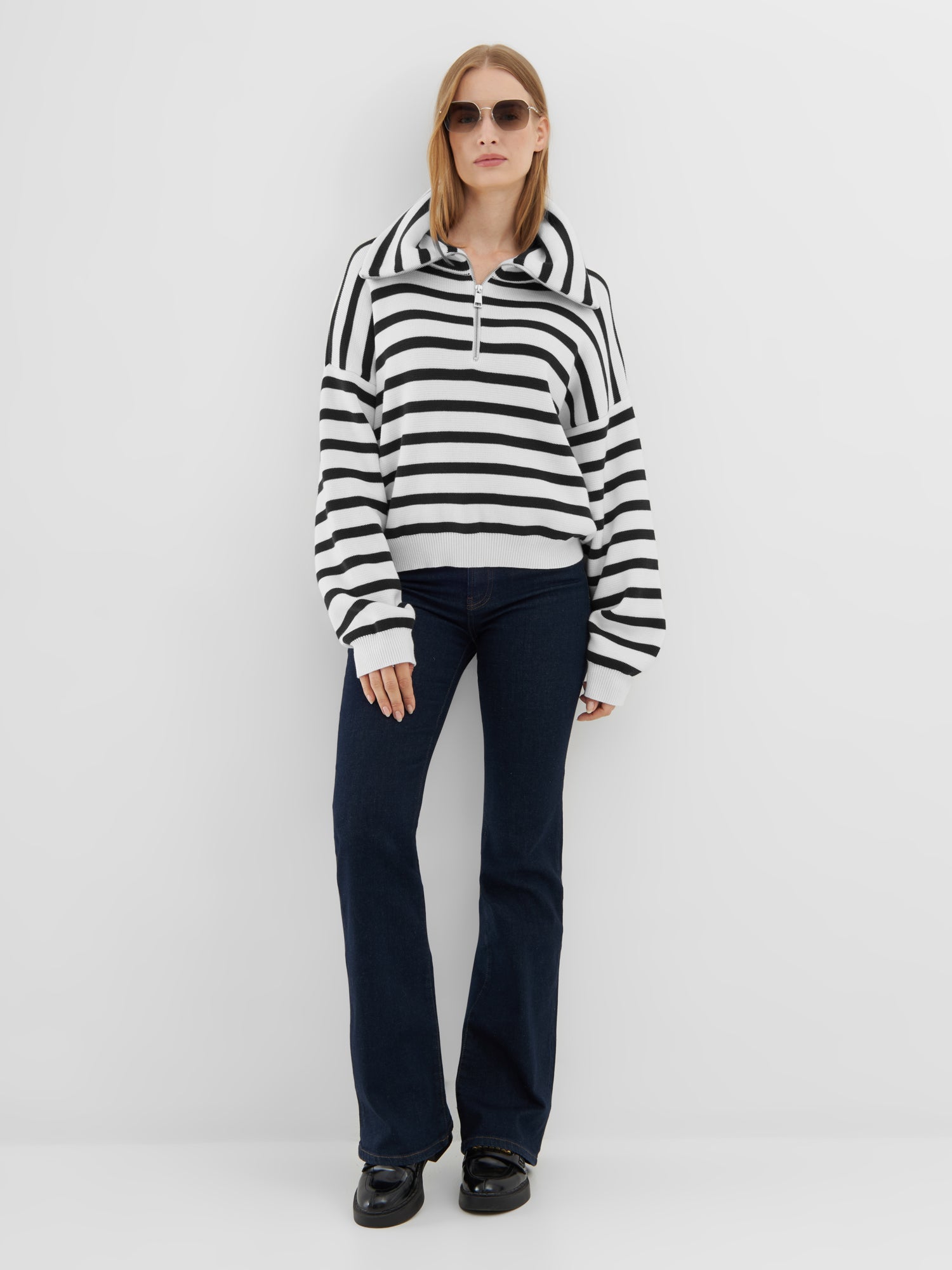 Eine Frau mit Sonnenbrille, dem Pullover Zipper Sailor, dunkelblauen Röhrenjeans und schwarzen Schuhen steht vor einem schlichten weißen Hintergrund.