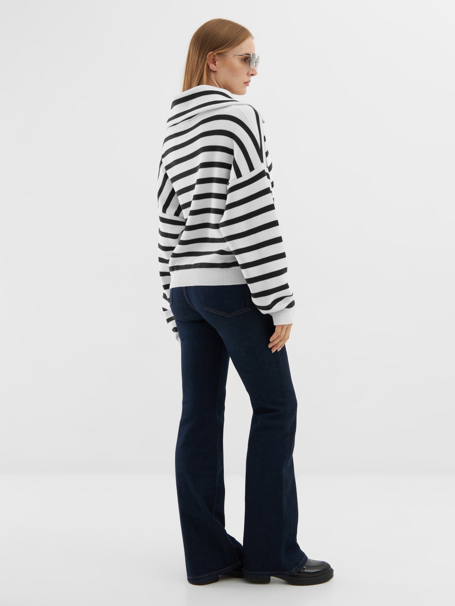 Eine Person, die eine Sonnenbrille, den Pullover Zipper Sailor, dunkle Schlagjeans und schwarze Schuhe trägt, steht vor einem schlichten weißen Hintergrund, schaut zur Seite und schaut weg.