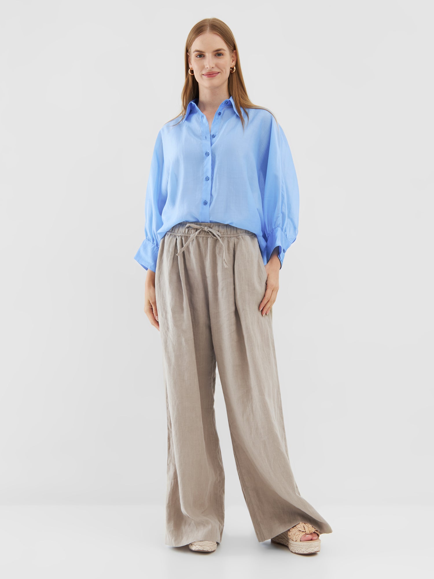 Eine Frau mit glattem, langem Haar lächelt vor einem schlichten Hintergrund in dem Bluse Antonia short Sleeve Blue - einem übergroßen hellblauen Button-up-Hemd - kombiniert mit einer hellbeigen, weit geschnittenen Hose und beigen Plateauschuhen.