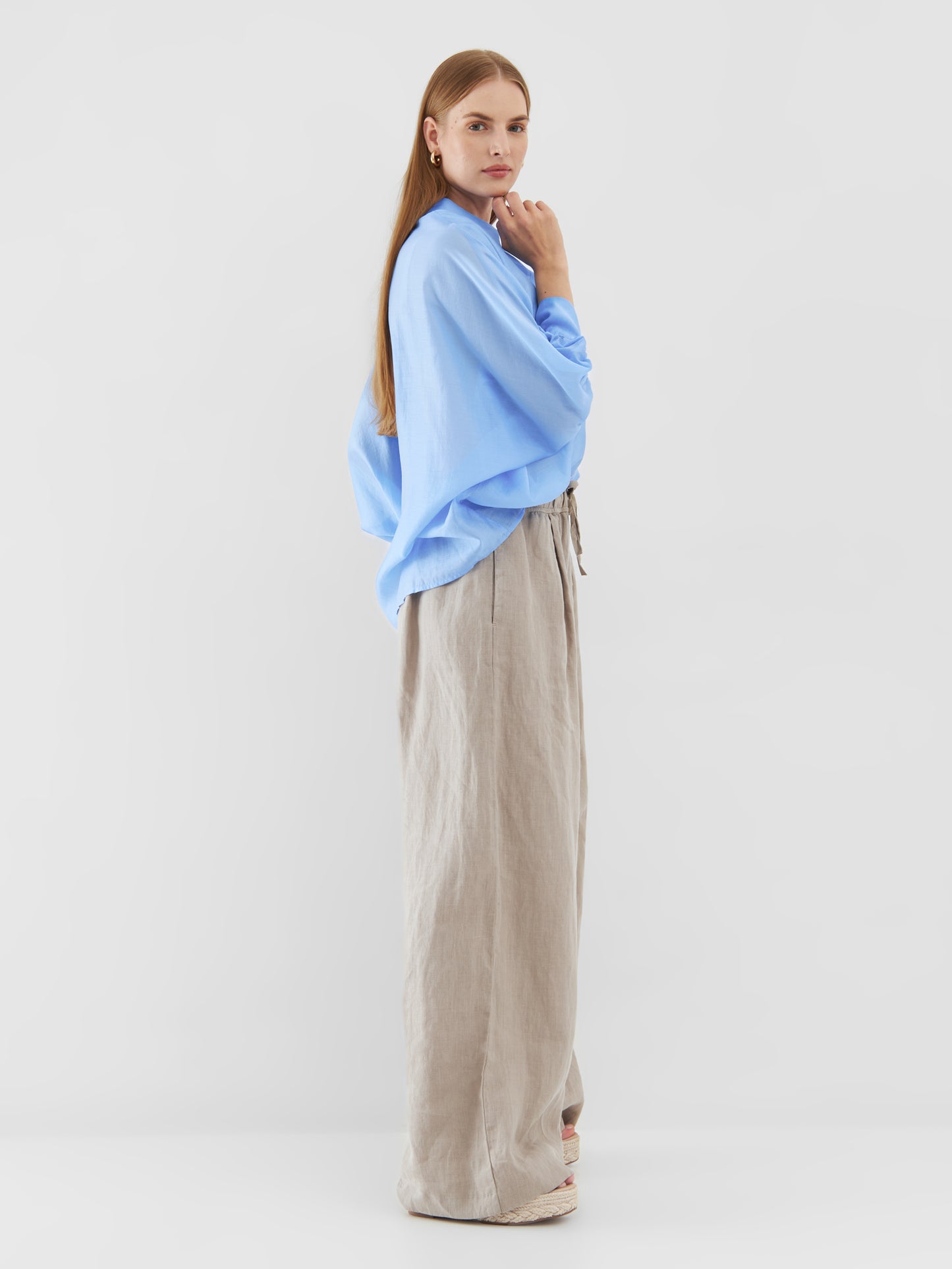 Eine Frau steht im Profil vor einem schlichten Hintergrund. Sie trägt ein übergroßes Bluse Antonia Short Sleeve Blue und eine beige Hose mit weitem Bein. Sie hat langes glattes Haar, einen aufmerksamen Gesichtsausdruck und eine Hand am Kinn erhoben.