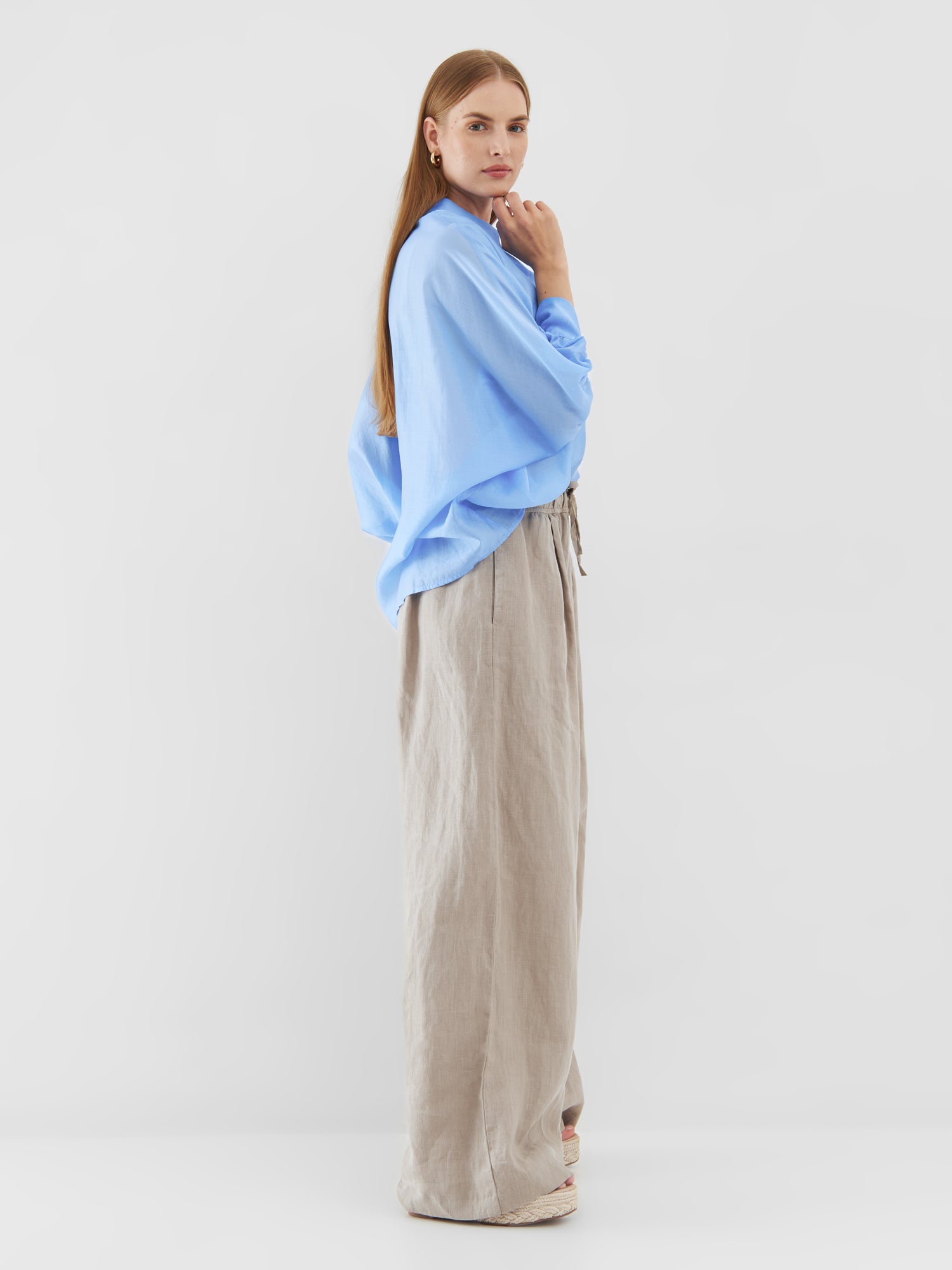 Eine Frau steht im Profil vor einem schlichten Hintergrund. Sie trägt ein übergroßes Bluse Antonia Short Sleeve Blue und eine beige Hose mit weitem Bein. Sie hat langes glattes Haar, einen aufmerksamen Gesichtsausdruck und eine Hand am Kinn erhoben.