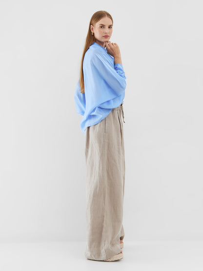 Eine Frau steht im Profil vor einem schlichten Hintergrund. Sie trägt ein übergroßes Bluse Antonia Short Sleeve Blue und eine beige Hose mit weitem Bein. Sie hat langes glattes Haar, einen aufmerksamen Gesichtsausdruck und eine Hand am Kinn erhoben.