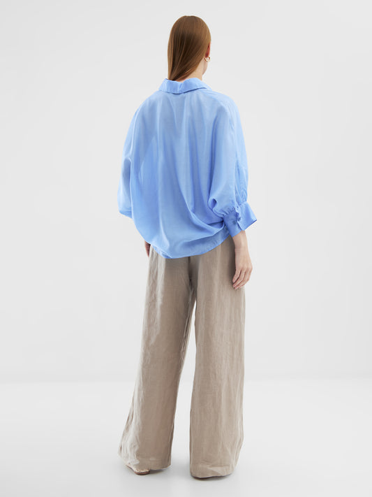 Eine Person mit glattem, langem Haar steht mit dem Gesicht zur Wand in einem minimalistischen weißen Raum und trägt eine übergroße Bluse Antonia Short Sleeve Blue und eine beigefarbene Hose mit weiten Beinen.