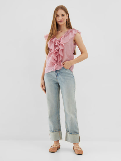 Eine Frau steht vor einem schlichten weißen Hintergrund, trägt das Top Ruffle Light Pink, hellblaue Jeans mit Bündchen und beigefarbene Sandalen, eine Hand in der Tasche und einen entspannten Gesichtsausdruck.