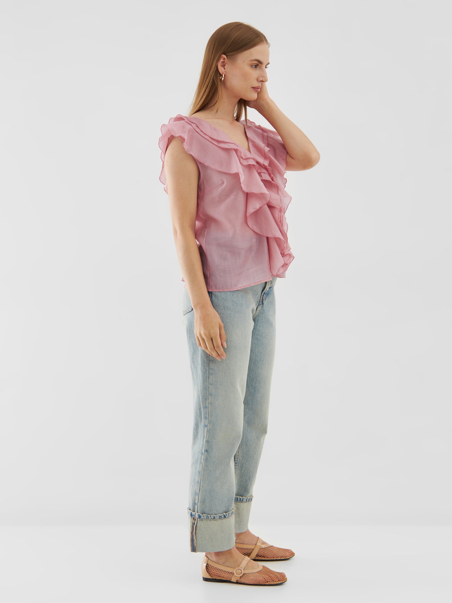 Eine Frau steht seitlich und trägt das Top Ruffle Light Pink, hellblaue, hochgekrempelte Jeans, rosa Webschuhe und Ohrringe, während sie mit einer Hand ihr Haar vor einem schlichten weißen Hintergrund berührt.