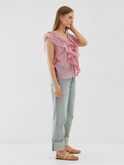 Eine Frau steht seitlich und trägt das Top Ruffle Light Pink, hellblaue, hochgekrempelte Jeans, rosa Webschuhe und Ohrringe, während sie mit einer Hand ihr Haar vor einem schlichten weißen Hintergrund berührt.