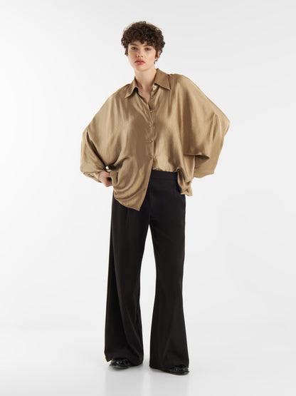 Eine Person mit kurzen lockigen Haaren steht selbstbewusst vor einem weißen Hintergrund, trägt die Bluse Antonia Shiny Beige mit weiten Ärmeln und eine schwarze Hose mit weitem Bein, eine Hand an der Hüfte.