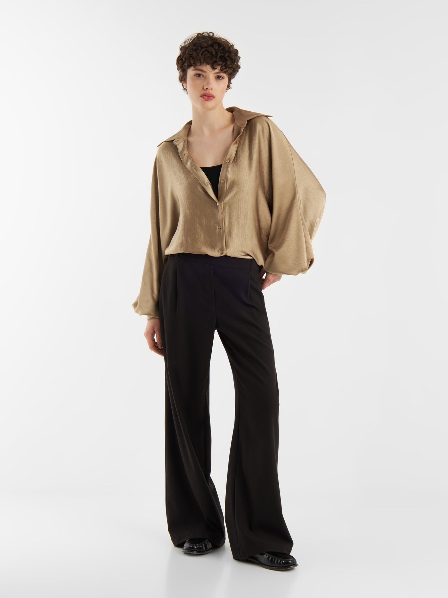 Eine Person mit kurzen lockigen Haaren posiert vor einem schlichten weißen Hintergrund und trägt eine übergroße Bluse Antonia Shiny Beige über einem schwarzen Oberteil, gepaart mit einer schwarzen Hose mit weitem Bein und schwarzen Schuhen.
