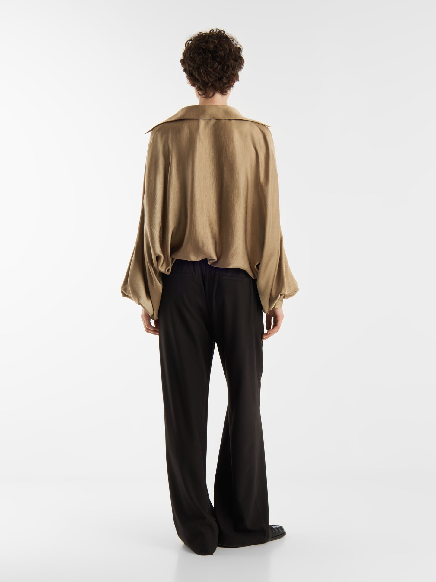 Eine Person mit kurzen lockigen Haaren, von hinten gesehen, trägt die Bluse Antonia Shiny Beige mit weiten Ärmeln und einer schwarzen Hose mit weitem Bein, die vor einem schlichten weißen Hintergrund steht.