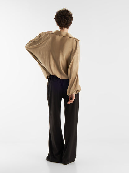 Eine Person mit kurzen lockigen Haaren steht mit dem Gesicht zur Wand, trägt eine übergroße Bluse Antonia Shiny Beige und eine schwarze Hose mit weitem Bein vor einem schlichten weißen Hintergrund. Ein Arm ist angewinkelt, die Hand ruht auf dem oberen Rücken.