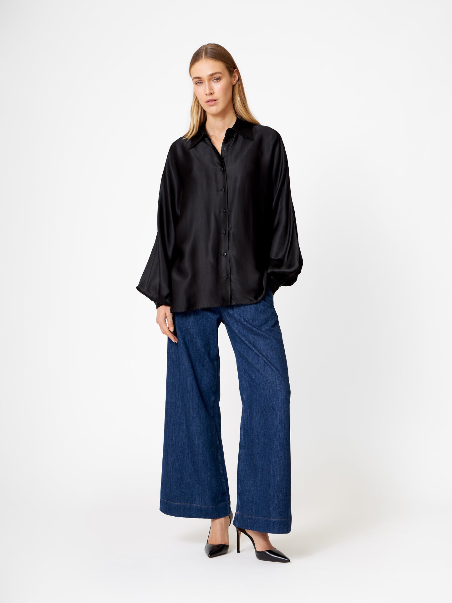 Eine Frau mit langem, glattem, blondem Haar und neutralem Gesichtsausdruck steht vor einem weißen Hintergrund. Sie trägt die Bluse Antonia Shiny Black in einer übergroßen Passform, Bluejeans mit weitem Bein und schwarze, spitz zulaufende Absätze.