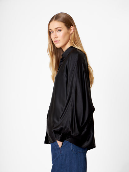 Eine Person mit langen blonden Haaren steht seitlich, trägt eine Bluse Antonia Shiny Black im Oversized-Look - eine lockere schwarze Satinbluse -, blaue Jeans und eine Hand in der Tasche, vor einem schlichten weißen Hintergrund.