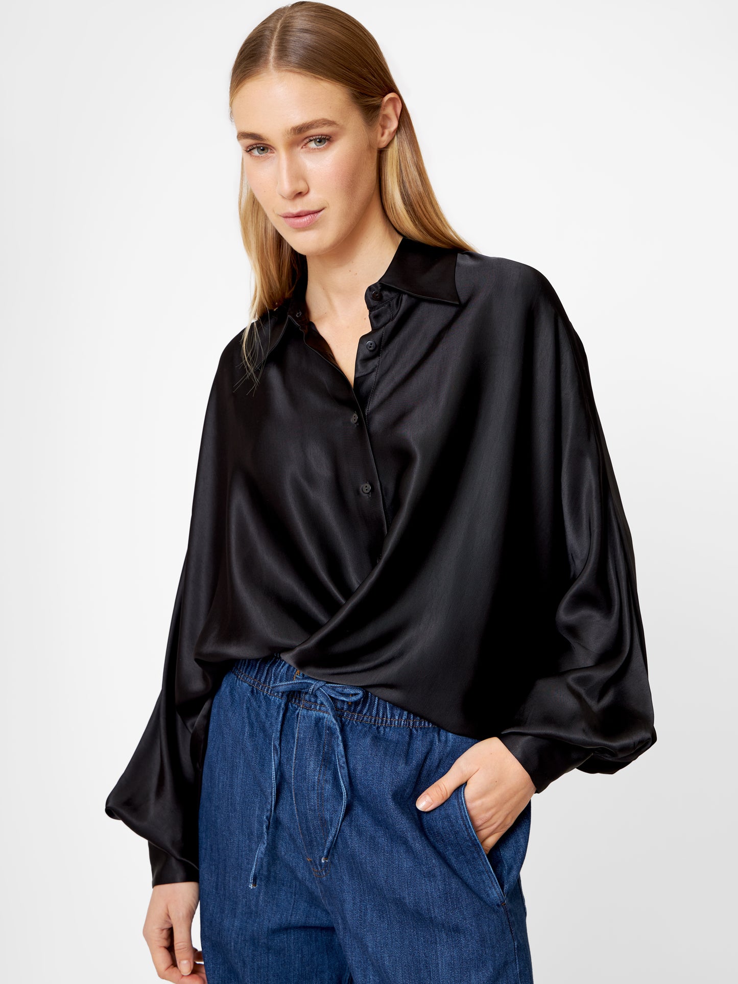 Eine Frau mit langen, glatten blonden Haaren trägt eine Bluse Antonia Shiny Black im Oversized-Look, eine locker sitzende Satinbluse, gepaart mit einer blauen Kordeljeans. Sie steht mit einer Hand in der Tasche vor einem schlichten weißen Hintergrund.