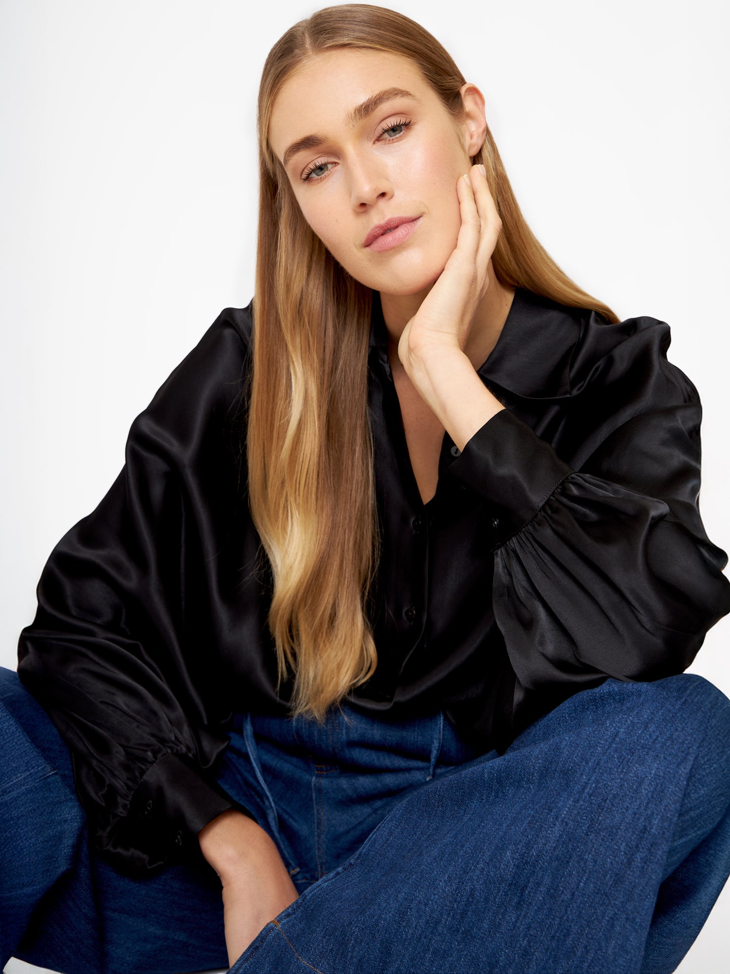 Eine Frau mit langen blonden Haaren sitzt lässig da und stützt ihren Kopf auf ihre Hand. Sie trägt die Bluse Antonia Shiny Black im Oversized-Look mit weiten Ärmeln und blauen Jeans vor einem schlichten weißen Hintergrund.