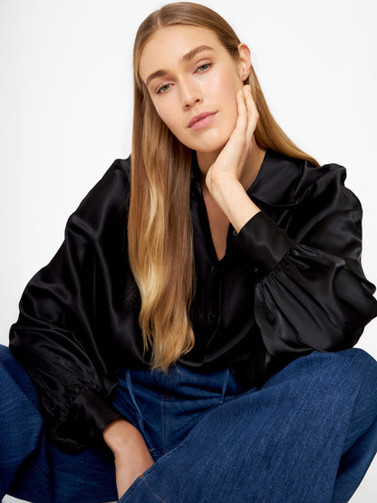 Eine Frau mit langen blonden Haaren sitzt lässig da und stützt ihren Kopf auf ihre Hand. Sie trägt die Bluse Antonia Shiny Black im Oversized-Look mit weiten Ärmeln und blauen Jeans vor einem schlichten weißen Hintergrund.