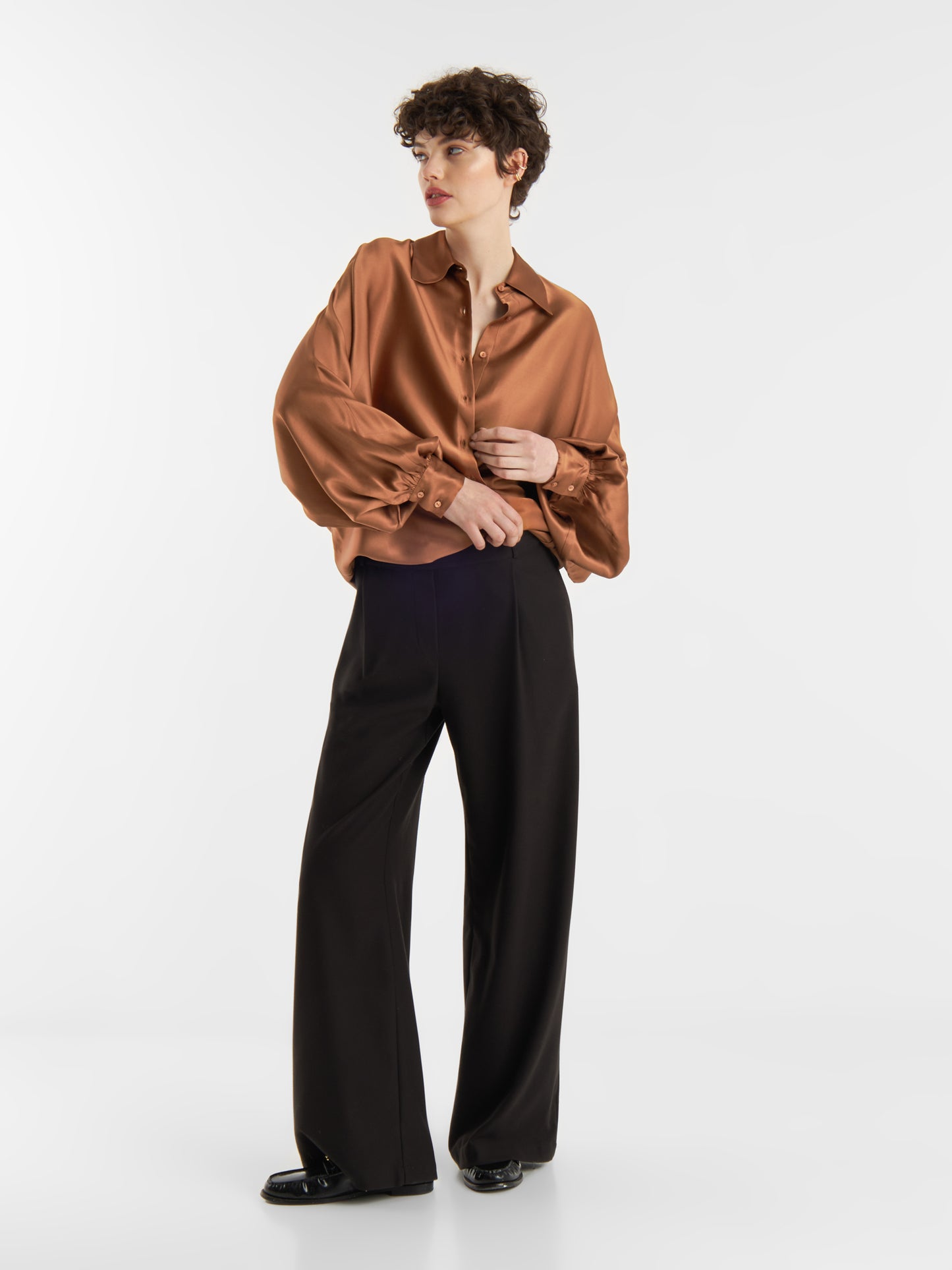 Eine Person mit kurzen lockigen Haaren modelliert die Bluse Antonia Shiny Brown im Oversized-Look und eine schwarze Hose mit weitem Bein. Sie steht in entspannter Pose vor einem schlichten weißen Hintergrund.