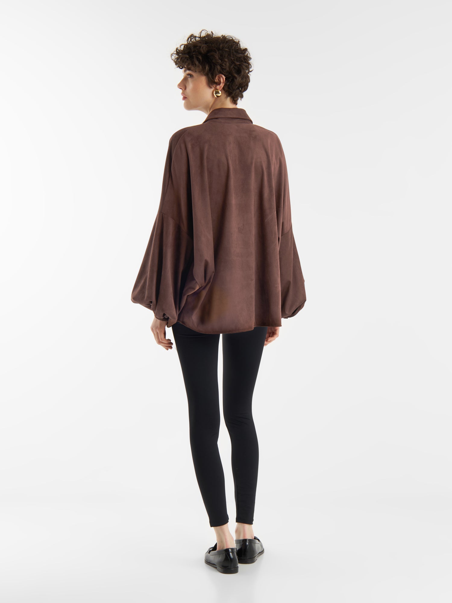 Eine Person mit kurzen lockigen Haaren, die die Bluse Antonia Vegan Leather Brown, schwarze Leggings und schwarze Schuhe trägt, steht mit dem Gesicht nach unten vor einem schlichten weißen Hintergrund.