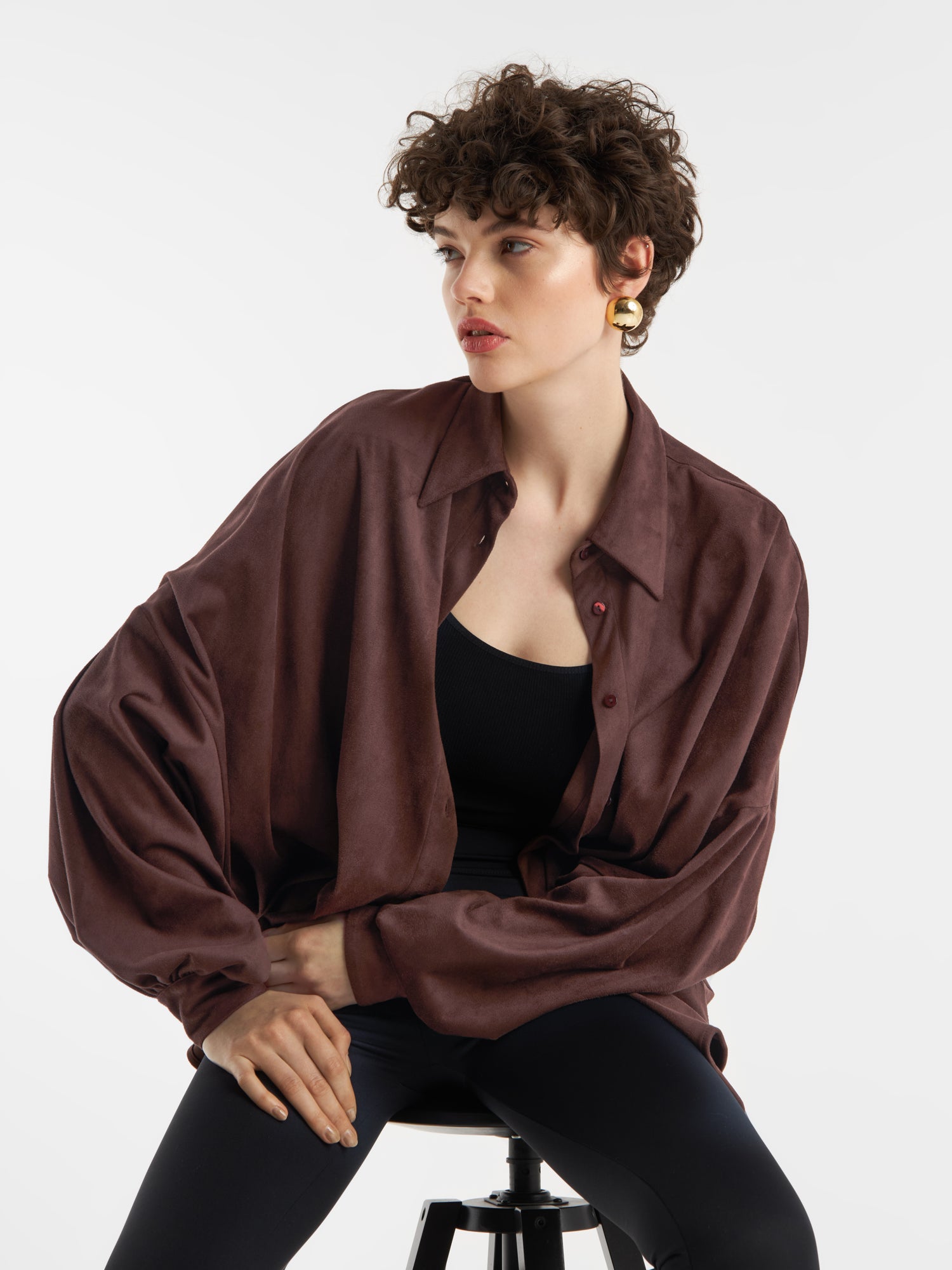 Eine Person mit kurzen lockigen Haaren, die die Bluse Antonia Vegan Leather Brown über einem schwarzen Oberteil und einer schwarzen Hose trägt, sitzt auf einem Hocker vor einem weißen Hintergrund. Sie trägt goldene Ohrringe und hat einen neutralen Gesichtsausdruck.