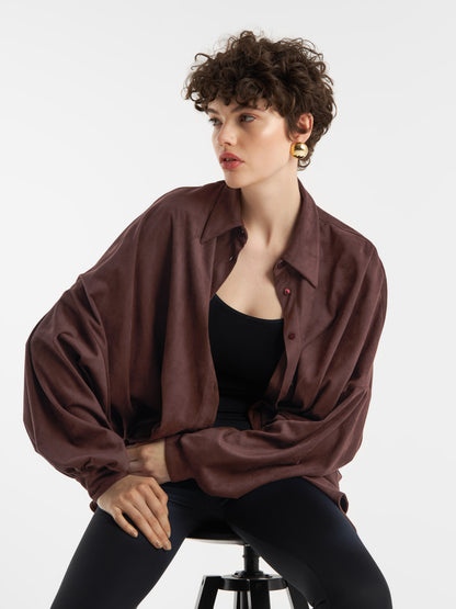 Eine Person mit kurzen lockigen Haaren, die die Bluse Antonia Vegan Leather Brown über einem schwarzen Oberteil und einer schwarzen Hose trägt, sitzt auf einem Hocker vor einem weißen Hintergrund. Sie trägt goldene Ohrringe und hat einen neutralen Gesichtsausdruck.