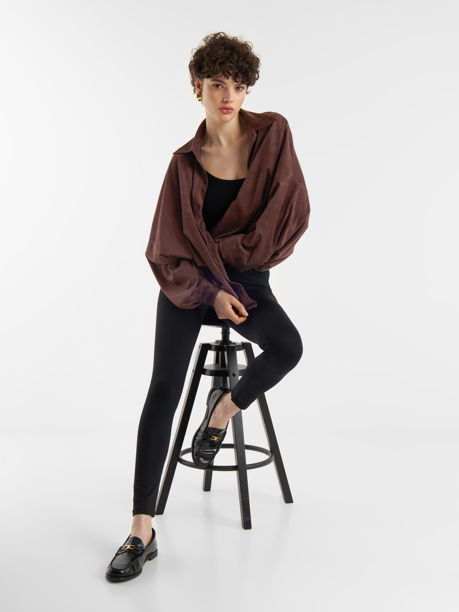 Eine Person mit kurzen lockigen Haaren sitzt auf einem schwarzen Hocker in einem Studio und trägt die Bluse Antonia Vegan Leather Brown über einem schwarzen Oberteil und Leggings mit schwarzen Halbschuhen vor einem schlichten weißen Hintergrund.