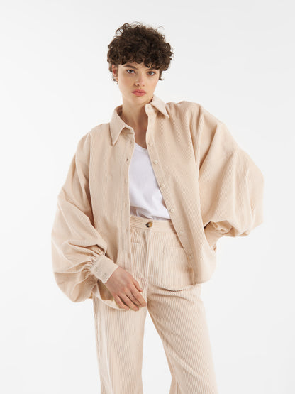 Eine Person mit kurzen, lockigen Haaren trägt die Bluse Antonia Corduroy OFW in Beige über einem weißen Oberteil und einer passenden Cordhose und steht vor einem einfarbig weißen Hintergrund.