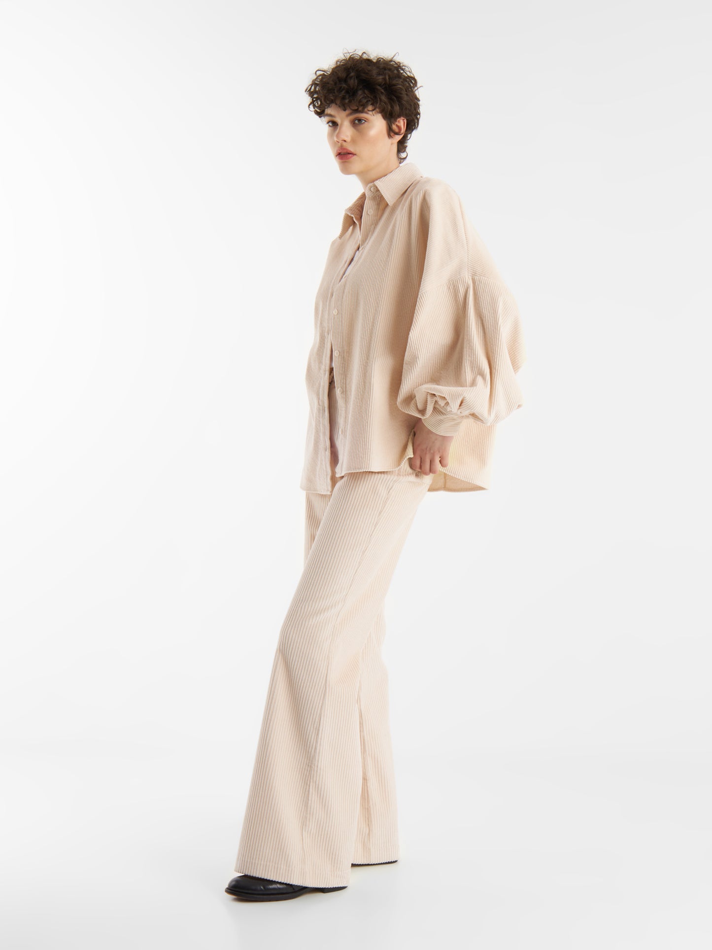 Eine Person mit kurzen lockigen Haaren trägt die Bluse Antonia Corduroy OFW und eine dazu passende Schlaghose. Sie steht vor einem schlichten weißen Hintergrund und schaut selbstbewusst zur Seite.