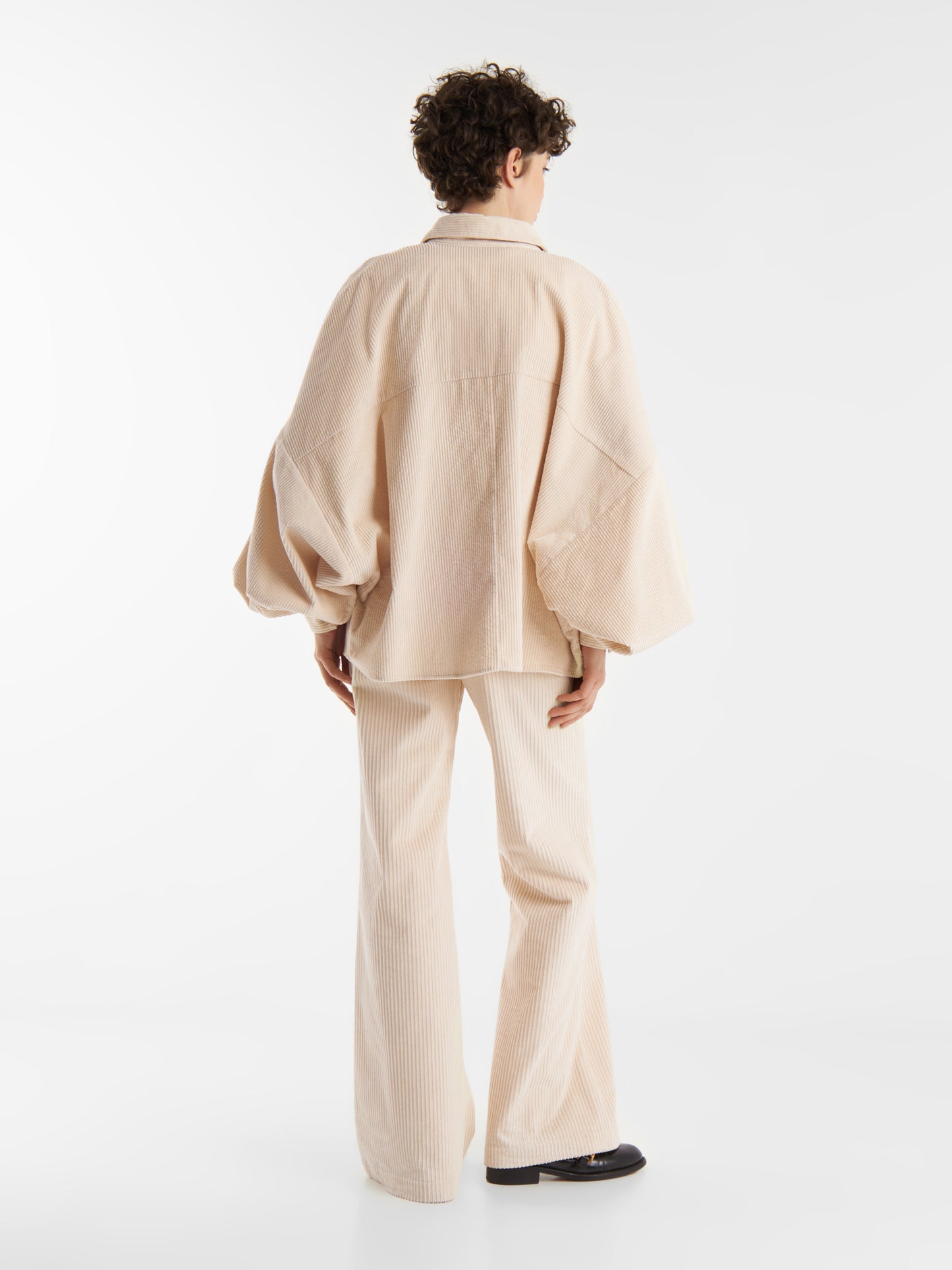 Eine Person mit kurzen lockigen Haaren blickt von sich weg und trägt die Bluse Antonia Corduroy OFW mit Ballonärmeln und passender Hose mit weitem Bein vor einem schlichten weißen Hintergrund.