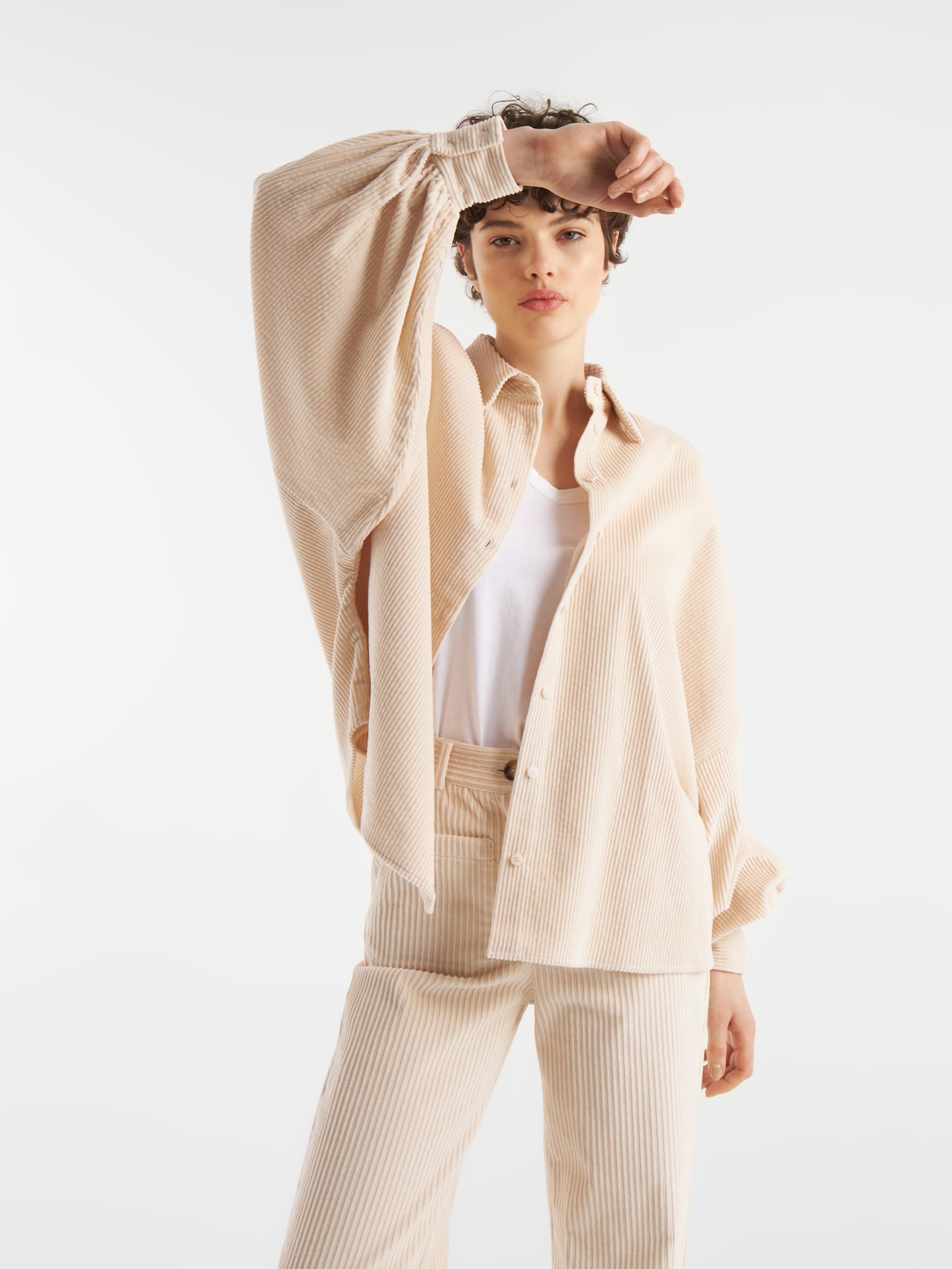 Eine Person mit kurzem, lockigem Haar trägt die Bluse Antonia Corduroy OFW in Beige über einem weißen Top und passenden Hosen, steht vor weißem Hintergrund und hebt einen Arm, um den Kopf zu berühren.