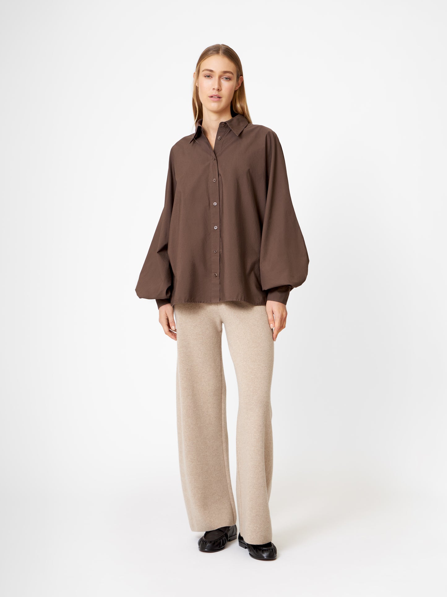 Eine Frau mit glattem hellbraunem Haar und einem neutralen Gesichtsausdruck steht vor einem weißen Hintergrund und trägt eine übergroße Bluse Antonia Espresso, eine beigefarbene Hose mit weitem Bein und schwarze Schuhe.