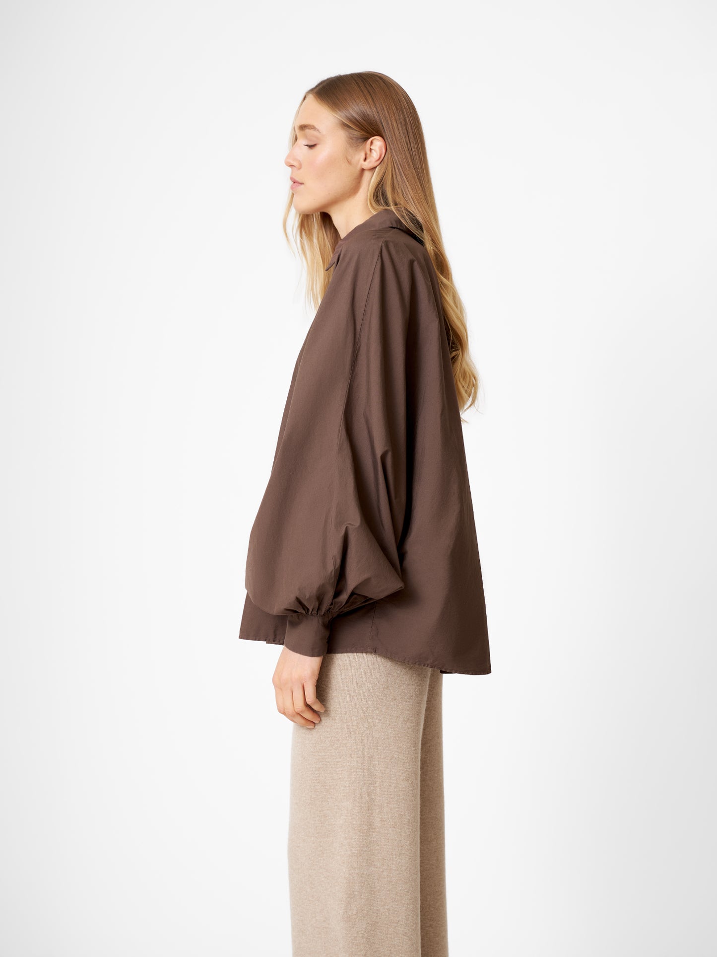 Eine Frau mit langen blonden Haaren steht im Profil vor einem unifarbenen Hintergrund und trägt die übergroße Bluse Antonia Espresso mit Fledermausärmeln und hellbeiger Hose.